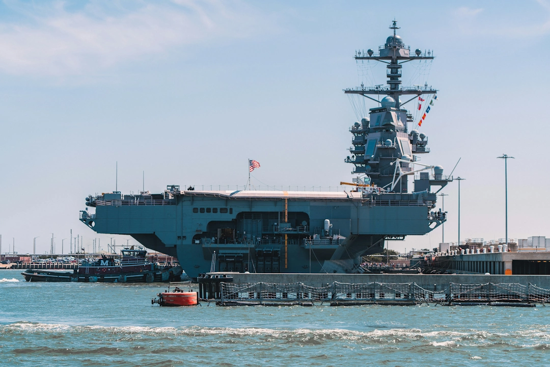 Docked aircraft carrier in Norfolk, Virginia naval port, with American flag flying and support vessels in background.